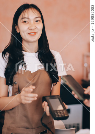 Asian senior female barista adult receiving mobile payment from customer using smartphone at cafe demonstrating modern cashless transaction in cozy local family coffee shop Asian senior female barista adult receiving mobile payment from customer using smartphone at cafe demonstrating modern cashless transaction in cozy local family coffee shop 139883082