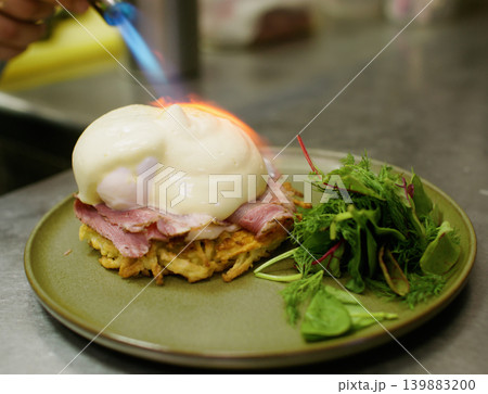 Preparing egg benedict. Healthy meal. Professional chef man hand using culinary torch burning salad ham fresh asparagus dish on plate in restaurant. Preparing egg benedict. Healthy meal. Professional chef man hand using culinary torch burning salad ham fresh asparagus dish on plate in restaurant. 139883200