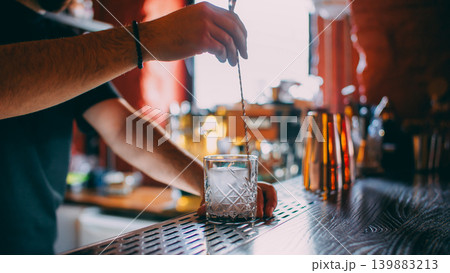 Cocktail making. Ice cube mixing. Unrecognizable barman hands stirring alcohol drink with professional spoon in transparent glass on bar counter. Cocktail making. Ice cube mixing. Unrecognizable barman hands stirring alcohol drink with professional spoon in transparent glass on bar counter. 139883213
