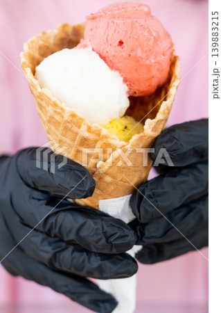 Serving dessert. Fruit scoop. Gelato flavor mix in crispy waffle cone. Pink white color sorbet ice cream ball in black glove hand. Serving dessert. Fruit scoop. Gelato flavor mix in crispy waffle cone. Pink white color sorbet ice cream ball in black glove hand. 139883215