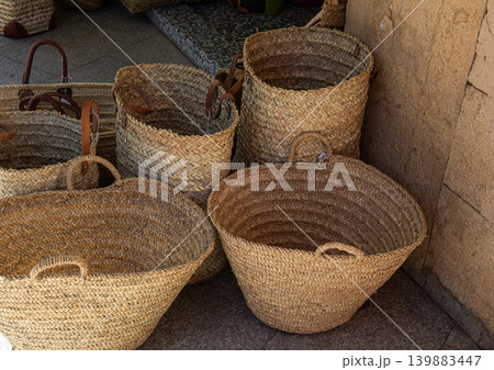 Different types of handmade wicker baskets and woven straw bags are arranged and stacked on a stone floor in a market area during daylight hours. Different types of handmade wicker baskets and woven straw bags are arranged and stacked on a stone floor in a market area during daylight hours. 139883447
