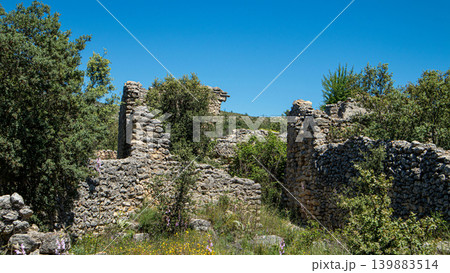 Stone buildings and ruins remain from an old settlement. The area is full of Mediterranean trees and wild flowers under a clear blue sky. Nature has reclaimed the land, offering an interesting view. Stone buildings and ruins remain from an old settlement. The area is full of Mediterranean trees and wild flowers under a clear blue sky. Nature has reclaimed the land, offering an interesting view. 139883514