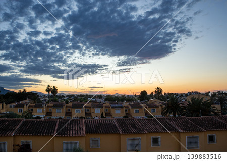 The sun sets over a Mediterranean town filled with terracotta rooftops. The sky is dramatic with clouds. This moment captures the end of a day in a lively location. The sun sets over a Mediterranean town filled with terracotta rooftops. The sky is dramatic with clouds. This moment captures the end of a day in a lively location. 139883516