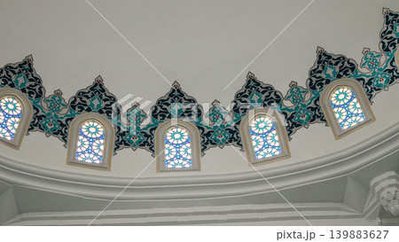 An elegant circular wall plaque with black Arabic script is displayed against a clean white background in a well-lit architectural setting. This space features decorative elements and windows. An elegant circular wall plaque with black Arabic script is displayed against a clean white background in a well-lit architectural setting. This space features decorative elements and windows. 139883627