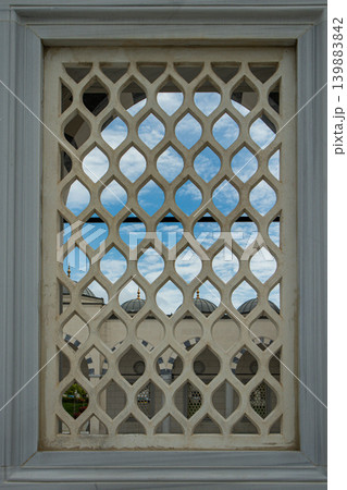 View shows mosque domes framed by a white stone lattice window. The sun is setting, casting shadows through the geometric design. The blue sky adds depth to the scene. View shows mosque domes framed by a white stone lattice window. The sun is setting, casting shadows through the geometric design. The blue sky adds depth to the scene. 139883842