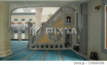 Inside a modern Islamic place of worship, a marble minbar shows golden carvings and intricate latticework. The space has blue carpets and large windows letting in light. Inside a modern Islamic place of worship, a marble minbar shows golden carvings and intricate latticework. The space has blue carpets and large windows letting in light. 139884166