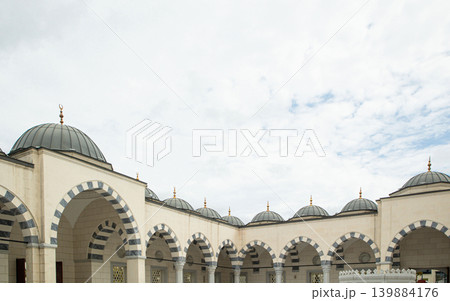 A mosque stands tall with a prominent minaret. Intricate stone patterns cover the exterior. Arched balconies add to the structure's design. Clouds drift above the scene. A mosque stands tall with a prominent minaret. Intricate stone patterns cover the exterior. Arched balconies add to the structure's design. Clouds drift above the scene. 139884176