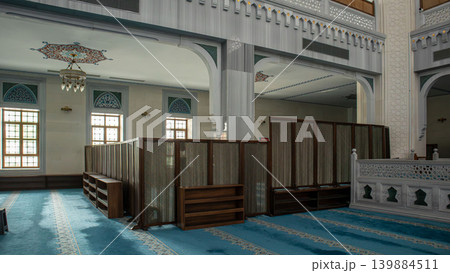 A mosque prayer area features teal carpeting and wooden privacy screens. Natural light streams through traditional windows, creating a space for prayer and reflection. A mosque prayer area features teal carpeting and wooden privacy screens. Natural light streams through traditional windows, creating a space for prayer and reflection. 139884511