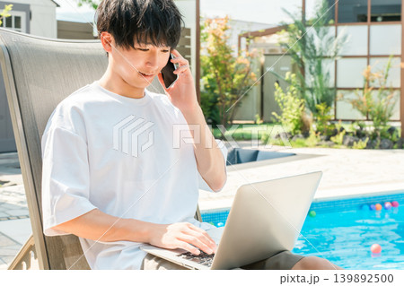 プールサイドのリクライニングチェアに座りスマホで通話,パソコンを操作する男性 プールサイドのリクライニングチェアに座りスマホで通話,パソコンを操作する男性 139892500
