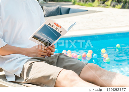プールサイドのリクライニングチェアに座りガイドブックを読む若いアジア人男性 プールサイドのリクライニングチェアに座りガイドブックを読む若いアジア人男性 139892577