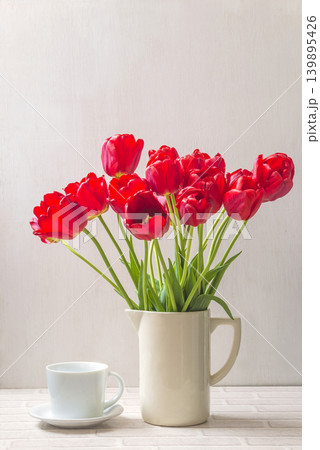 Bouquet of red tulip flowers in the white jug and cup of tea or coffee on a table against white background 139895426