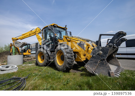A yellow backhoe works at a construction site during the day. Another backhoe is visible in the background. The site has various construction materials. loader A yellow backhoe works at a construction site during the day. Another backhoe is visible in the background. The site has various construction materials. loader 139897536
