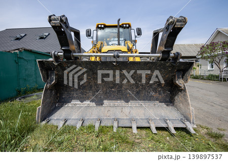 A large yellow loader is parked on a construction site with grass and buildings visible under a bright blue sky during daytime A large yellow loader is parked on a construction site with grass and buildings visible under a bright blue sky during daytime 139897537