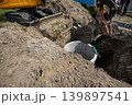 A construction crew is digging a hole to install a sewer system. A machine is lifting a concrete pipe at a site. The sun is shining in the afternoon 139897541