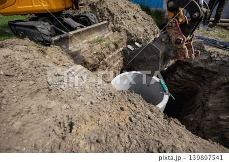 A construction crew is digging a hole to install a sewer system. A machine is lifting a concrete pipe at a site. The sun is shining in the afternoon A construction crew is digging a hole to install a sewer system. A machine is lifting a concrete pipe at a site. The sun is shining in the afternoon 139897541