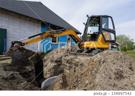 A digger is at work digging a hole beside a structure. The site shows signs of construction with dirt and machinery present on a clear day A digger is at work digging a hole beside a structure. The site shows signs of construction with dirt and machinery present on a clear day 139897542