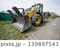 A large construction machine sits on the side of a dirt road close to houses. The machine has a bucket in front and yellow paint on its body. loader 139897543
