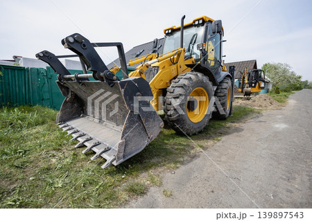 A large construction machine sits on the side of a dirt road close to houses. The machine has a bucket in front and yellow paint on its body. loader 139897543