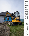 An excavator is digging near a house with blue walls while workers plan for an ongoing renovation project in the daytime 139897545