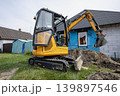 A small excavator is digging in front of a house. The background shows a blue tarp on the wall and a small shed nearby. Grass surrounds the area 139897546