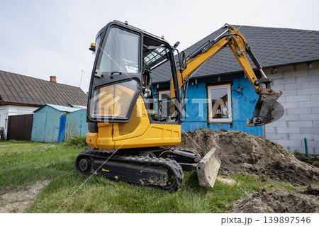 A small excavator is digging in front of a house. The background shows a blue tarp on the wall and a small shed nearby. Grass surrounds the area 139897546