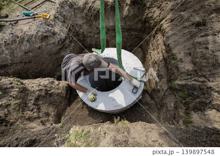 A worker lowers a concrete round structure into a hole at a construction site during daytime. Tools and dirt are present around the worker A worker lowers a concrete round structure into a hole at a construction site during daytime. Tools and dirt are present around the worker 139897548