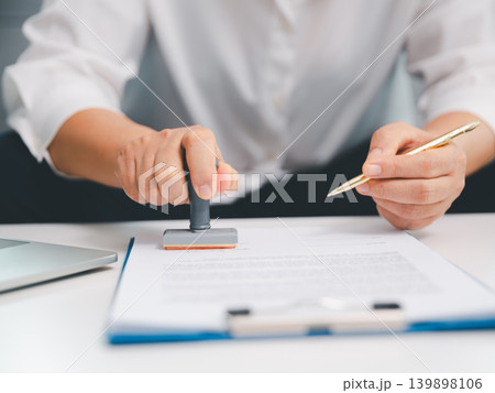 Professional hand pressing rubber stamp on business document while holding pen, showing approval, certification, legal paperwork, contract verification, finance process, and office workflow. Professional hand pressing rubber stamp on business document while holding pen, showing approval, certification, legal paperwork, contract verification, finance process, and office workflow. 139898106