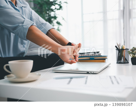 Professional stretching hands after closing laptop on white office desk with coffee cup, notebooks, and reports, showing work break, wellness, productivity, stress relief, and modern workspace 139898107