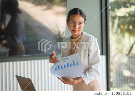 Professional woman in white blazer presenting sales report and bar chart near laptop, using confident gesture for business meeting, finance review, marketing strategy, and office teamwork. Professional woman in white blazer presenting sales report and bar chart near laptop, using confident gesture for business meeting, finance review, marketing strategy, and office teamwork. 139898108