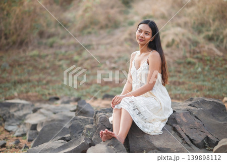 Woman in white lace dress sitting on dark rocks, looking thoughtful in soft natural light, peaceful outdoor portrait, bohemian summer fashion, wellness lifestyle, relaxation, travel, Woman in white lace dress sitting on dark rocks, looking thoughtful in soft natural light, peaceful outdoor portrait, bohemian summer fashion, wellness lifestyle, relaxation, travel, 139898112