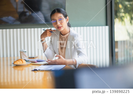 Professional businesswoman pointing to notes at an office table with pen, coffee, croissant, and paperwork, presenting planning details, business tasks, collaboration, and productive meeting workflow. 139898225