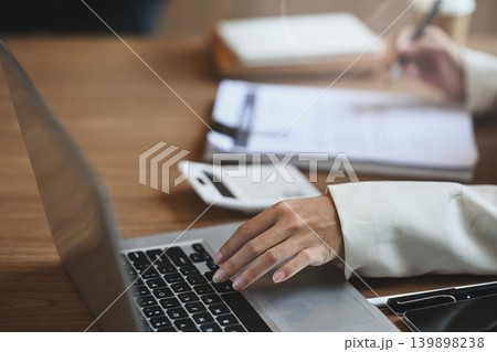 Businesswoman typing on a laptop at a desk with stack of papers and calculator Businesswoman typing on a laptop at a desk with stack of papers and calculator 139898238