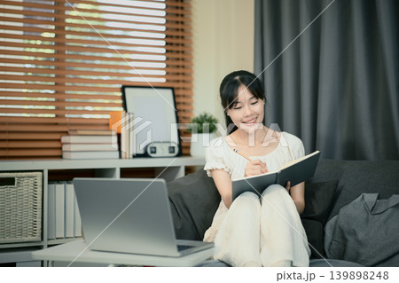 Smiling young woman remotely, sitting on sofa with notebooks and laptop, taking notes Smiling young woman remotely, sitting on sofa with notebooks and laptop, taking notes 139898248