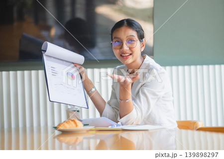 Confident businesswoman smiling while presenting business report charts in bright office, gesturing with open hand for financial data review, marketing strategy, and team briefing. Confident businesswoman smiling while presenting business report charts in bright office, gesturing with open hand for financial data review, marketing strategy, and team briefing. 139898297
