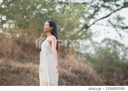 Woman in white lace dress standing in profile under green trees, breathing calmly in soft outdoor light, peaceful nature lifestyle, summer fashion, wellness, relaxation, freedom, and travel. Woman in white lace dress standing in profile under green trees, breathing calmly in soft outdoor light, peaceful nature lifestyle, summer fashion, wellness, relaxation, freedom, and travel. 139898300