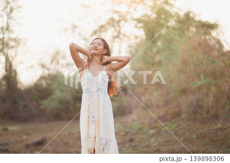 Woman in white lace dress stretching with hands behind neck, eyes closed in warm outdoor light, peaceful nature lifestyle portrait for wellness, self care, bohemian summer fashion, freedom, and joy. Woman in white lace dress stretching with hands behind neck, eyes closed in warm outdoor light, peaceful nature lifestyle portrait for wellness, self care, bohemian summer fashion, freedom, and joy. 139898306