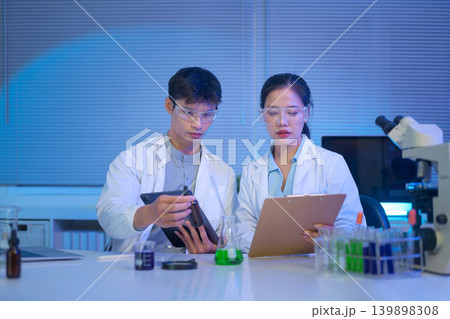 Two scientists discuss and record experimental data using tablet and clipboard in a modern laboratory with liquid samples and equipment Two scientists discuss and record experimental data using tablet and clipboard in a modern laboratory with liquid samples and equipment 139898308