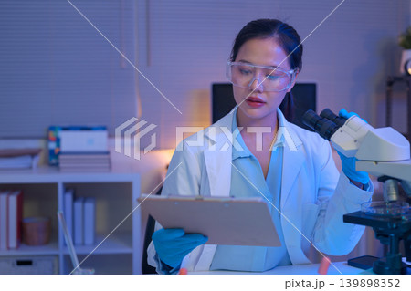 Female scientist using a microscope to analyze samples and writing on document Female scientist using a microscope to analyze samples and writing on document 139898352
