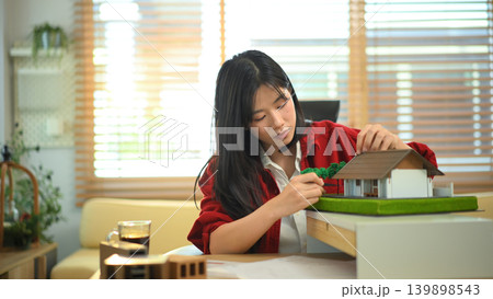 Female architect examining house model at office desk, Professional architect reviewing architectural house model Female architect examining house model at office desk, Professional architect reviewing architectural house model 139898543