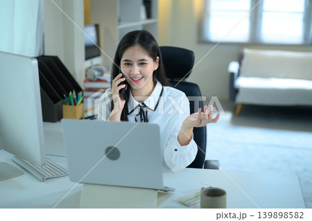 Young Asian businesswoman multitasking at a office desk, talking on a smartphone and using a laptop 139898582