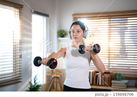 Focused woman performing dumbbell shoulder exercise indoors in bright modern home environment Focused woman performing dumbbell shoulder exercise indoors in bright modern home environment 139898706