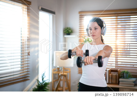 Focused woman performing dumbbell shoulder exercise indoors in bright modern home environment Focused woman performing dumbbell shoulder exercise indoors in bright modern home environment 139898714