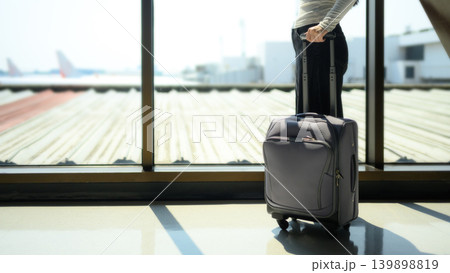Closeup young woman traveler waiting with suitcase watching airplane at airport window Closeup young woman traveler waiting with suitcase watching airplane at airport window 139898819