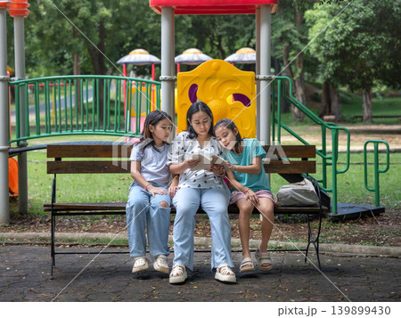Asian mother reading a book to her two young daughters while sitting on a bench in the park. 139899430