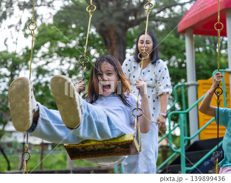 Caring Asian mother pushing her young daughter on a swing at the outdoor playground. Caring Asian mother pushing her young daughter on a swing at the outdoor playground. 139899436