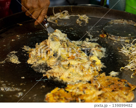 Close-up of a vendor cooking a traditional Thai crispy oyster omelet (Hoi Tod) on a large flat griddle. Close-up of a vendor cooking a traditional Thai crispy oyster omelet (Hoi Tod) on a large flat griddle. 139899482