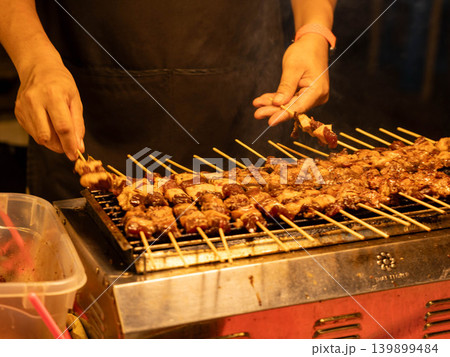 Close-up of a vendor grilling meat skewers on an electric grill at a street food market. Close-up of a vendor grilling meat skewers on an electric grill at a street food market. 139899484