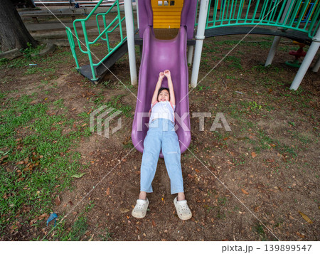 Cute young Asian girl having fun sliding down a purple slide at the outdoor playground. Cute young Asian girl having fun sliding down a purple slide at the outdoor playground. 139899547