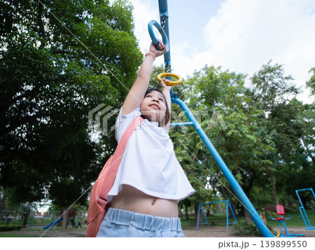 Low angle view of a brave young Asian girl hanging on a monkey bar at the outdoor playground. 139899550