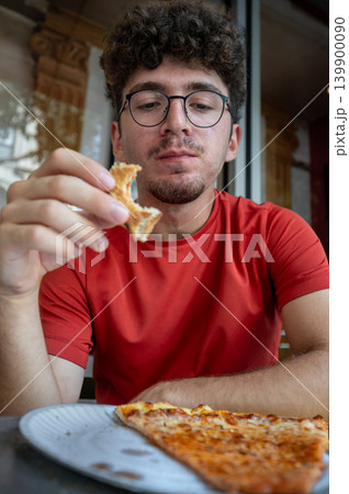 New york, usa august 19,2024. Young man in red shirt eats a new york pizza slice from a paper plate, enjoying a casual downtown lunch with focused bite 139900090